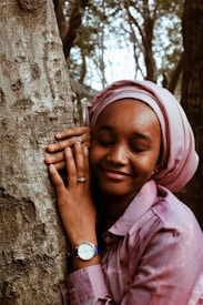 A person with a serene expression is embracing a tree trunk, wearing a pink headscarf and shirt. The watch and ring on their left hand suggest a sense of personal style. The background features a forest setting, with trees and diffused light creating a peaceful ambiance.