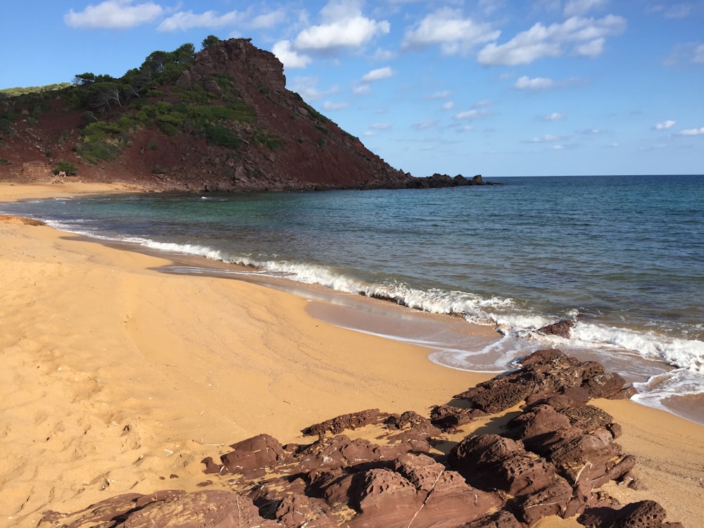 brown sand beach with green mountain in distance under blue sky and white clouds during daytime