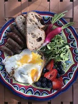 A colorful plate of food featuring fried eggs with runny yolks, slices of multigrain bread, grilled sausages, radishes with greens, fresh strawberries, blueberries, and a small pile of microgreens. The plate has a vibrant design with bold patterns and colors.