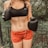 A person wearing a black sports bra and orange shorts is standing outdoors. They are holding up black boxing gloves, with a background of greenery and a metal fence.