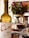 A bottle of Cune Rioja Crianza 2016 wine is placed prominently beside a filled wine glass. In the background, a cake decorated with marshmallows sits on a table covered with a decorative cloth. A colorful bouquet of roses and greenery adds a touch of elegance and vibrancy to the setting.