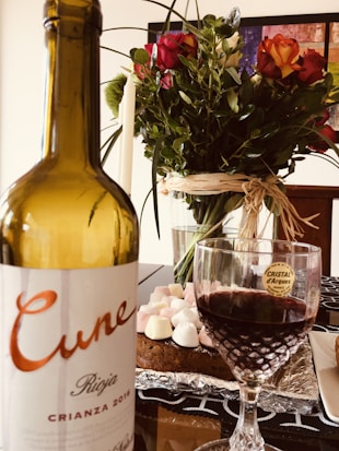 A bottle of Cune Rioja Crianza 2016 wine is placed prominently beside a filled wine glass. In the background, a cake decorated with marshmallows sits on a table covered with a decorative cloth. A colorful bouquet of roses and greenery adds a touch of elegance and vibrancy to the setting.