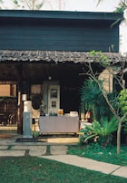 A quaint outdoor setup with a rustic wooden structure at the back, featuring a refreshment table adorned with a tablecloth and an array of items including jugs of orange juice and a water dispenser. The foreground is a neatly manicured lawn with a stone path leading to the table, flanked by lush greenery and various plants.