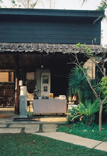 A quaint outdoor setup with a rustic wooden structure at the back, featuring a refreshment table adorned with a tablecloth and an array of items including jugs of orange juice and a water dispenser. The foreground is a neatly manicured lawn with a stone path leading to the table, flanked by lush greenery and various plants.