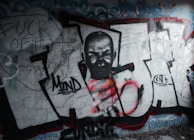 A graffiti wall features a monochrome portrait of a person with a fiercely intense expression, integrated into large letters outlined in black, with patches of red and orange spray paint. Surrounding the central art are scribbled words and rough sketches in darker tones. The overall composition appears rebellious and chaotic.