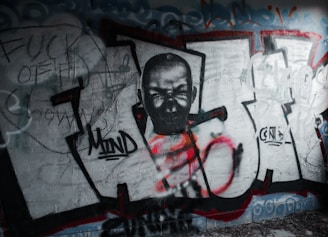 A graffiti wall features a monochrome portrait of a person with a fiercely intense expression, integrated into large letters outlined in black, with patches of red and orange spray paint. Surrounding the central art are scribbled words and rough sketches in darker tones. The overall composition appears rebellious and chaotic.