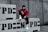 A person wearing a red jacket, black pants, and white sneakers is sitting on concrete barriers with 'NYPD' stenciled on them. The individual is also wearing a face mask and hoodie, giving a sense of anonymity. The background features a gray concrete wall.