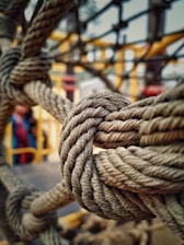 A close-up view of a thick rope knotted in an intricate pattern. The background is blurred, showcasing hints of a playground structure with yellow bars and a person wearing colorful clothes.