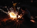 A person is using a power tool to cut metals, creating bright, intense sparks in a dimly lit environment. The individual is wearing a sleeveless shirt and seems focused on the task. Various wooden objects and metal pieces are scattered around.