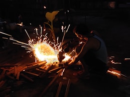 A person is using a power tool to cut metals, creating bright, intense sparks in a dimly lit environment. The individual is wearing a sleeveless shirt and seems focused on the task. Various wooden objects and metal pieces are scattered around.