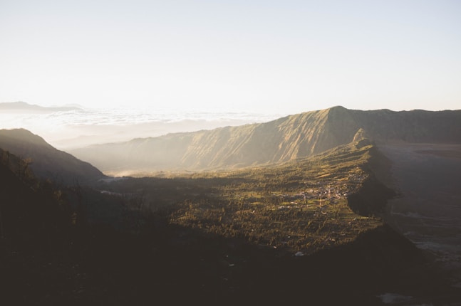 A scenic view of a mountainous landscape bathed in soft morning light, with a valley and scattered settlements below. The mountains have lush greenery on their slopes and are partially cloaked in mist, while the sky is clear above.