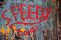 Bold red graffiti text spelling 'SPEEDY' is painted on a weathered and peeling blue metal surface with rust accents. The background shows patches of orange and yellow paint, adding a vibrant contrast to the scene.