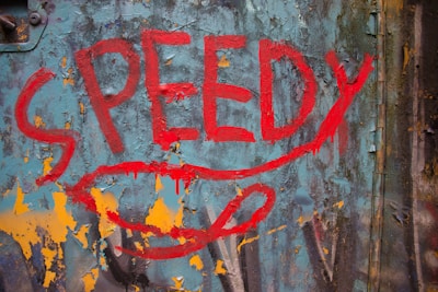 Bold red graffiti text spelling 'SPEEDY' is painted on a weathered and peeling blue metal surface with rust accents. The background shows patches of orange and yellow paint, adding a vibrant contrast to the scene.