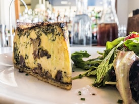 A slice of quiche featuring a rich, golden-brown crust and a filling packed with spinach and mushrooms is prominently placed on a white plate. Beside the quiche, a fresh green salad with mixed leaves is garnished with vinaigrette. In the background, a bar area with various bottles is slightly out of focus, adding to the setting's ambiance.