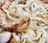 Close-up view of creamy fettuccine Alfredo with seasoned grilled chicken pieces. The pasta is richly coated in a white sauce, with visible herbs sprinkled throughout.