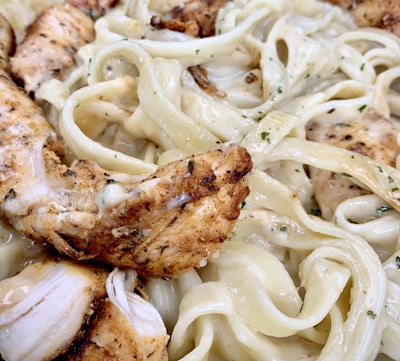 Close-up view of creamy fettuccine Alfredo with seasoned grilled chicken pieces. The pasta is richly coated in a white sauce, with visible herbs sprinkled throughout.