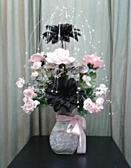 A floral arrangement with pink and black flowers in a clear vase filled with decorative crystals, adorned with a pink ribbon. The background features a neutral curtain, providing a soft backdrop for the vibrant flowers.