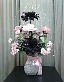 A floral arrangement with pink and black flowers in a clear vase filled with decorative crystals, adorned with a pink ribbon. The background features a neutral curtain, providing a soft backdrop for the vibrant flowers.