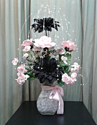 A floral arrangement with pink and black flowers in a clear vase filled with decorative crystals, adorned with a pink ribbon. The background features a neutral curtain, providing a soft backdrop for the vibrant flowers.