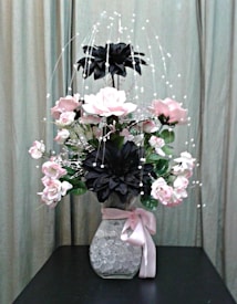 A floral arrangement with pink and black flowers in a clear vase filled with decorative crystals, adorned with a pink ribbon. The background features a neutral curtain, providing a soft backdrop for the vibrant flowers.