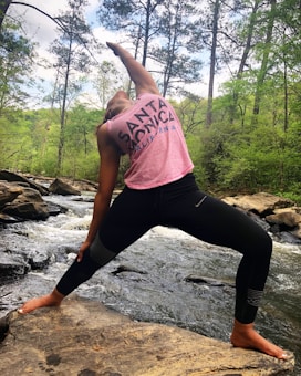 A person is performing a yoga pose on a large rock by a flowing river, surrounded by a lush forest with tall trees. The individual is wearing a pink tank top with 'Santa Monica, California' written on it and black leggings. The weather appears to be clear with a mix of sun and clouds visible in the sky.