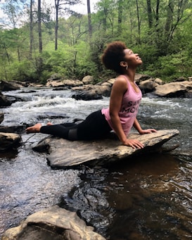 A person is practicing yoga on a large rock in the middle of a flowing stream. They are wearing a pink tank top and black leggings, stretching with their head tilted upwards. The surrounding environment is lush with green forest and trees, and rocks are scattered along the stream.