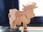 A wooden sculpture of a cow crafted with simple lines and shapes. The cow features a carved face with a neutral expression, two ears, and a tail made from a braided rope. It stands on a wooden surface against a dark wooden paneled background, with light coming from a window behind it.