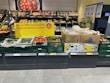 A supermarket display of various fresh produce including cucumbers, tomatoes, and packaged goods. The display is accompanied by a promotional sign in German indicating up to 100 unpackaged fruit and vegetable items. The fresh products are organized in green crates and cardboard boxes, with some wrapped in plastic. Price labels are visible at the bottom of each section.