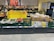 A supermarket display of various fresh produce including cucumbers, tomatoes, and packaged goods. The display is accompanied by a promotional sign in German indicating up to 100 unpackaged fruit and vegetable items. The fresh products are organized in green crates and cardboard boxes, with some wrapped in plastic. Price labels are visible at the bottom of each section.