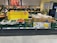 A supermarket display of various fresh produce including cucumbers, tomatoes, and packaged goods. The display is accompanied by a promotional sign in German indicating up to 100 unpackaged fruit and vegetable items. The fresh products are organized in green crates and cardboard boxes, with some wrapped in plastic. Price labels are visible at the bottom of each section.