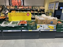 A supermarket display of various fresh produce including cucumbers, tomatoes, and packaged goods. The display is accompanied by a promotional sign in German indicating up to 100 unpackaged fruit and vegetable items. The fresh products are organized in green crates and cardboard boxes, with some wrapped in plastic. Price labels are visible at the bottom of each section.