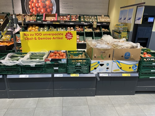A supermarket display of various fresh produce including cucumbers, tomatoes, and packaged goods. The display is accompanied by a promotional sign in German indicating up to 100 unpackaged fruit and vegetable items. The fresh products are organized in green crates and cardboard boxes, with some wrapped in plastic. Price labels are visible at the bottom of each section.