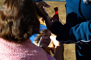 A person is having their hair cut or styled outdoors. They are looking into a handheld mirror to view the process. The person has a towel or cape draped around their shoulders, suggesting a hair styling session. The scene is brightly lit by natural sunlight.