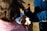 A person is having their hair cut or styled outdoors. They are looking into a handheld mirror to view the process. The person has a towel or cape draped around their shoulders, suggesting a hair styling session. The scene is brightly lit by natural sunlight.