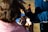 A person is having their hair cut or styled outdoors. They are looking into a handheld mirror to view the process. The person has a towel or cape draped around their shoulders, suggesting a hair styling session. The scene is brightly lit by natural sunlight.
