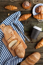 A rustic wooden table is adorned with a variety of pastries and breads. Sliced bread is placed on a cutting board over a striped blue and white cloth. Several croissants and cinnamon rolls, some on a decorative plate, are scattered around. There is a closed coffee cup and coffee beans sprinkled across the table, accompanied by a knife.