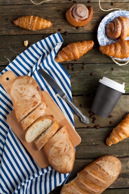 A rustic wooden table is adorned with a variety of pastries and breads. Sliced bread is placed on a cutting board over a striped blue and white cloth. Several croissants and cinnamon rolls, some on a decorative plate, are scattered around. There is a closed coffee cup and coffee beans sprinkled across the table, accompanied by a knife.