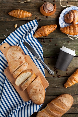 A rustic wooden table is adorned with a variety of pastries and breads. Sliced bread is placed on a cutting board over a striped blue and white cloth. Several croissants and cinnamon rolls, some on a decorative plate, are scattered around. There is a closed coffee cup and coffee beans sprinkled across the table, accompanied by a knife.