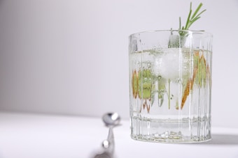 A clear glass of a refreshing cocktail garnished with rosemary, ice cubes, and slices of citrus. The background is minimalistic and bright, with a cocktail stirrer lying blurred in the foreground.