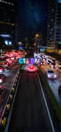 A busy urban street scene at night with heavy traffic, flanked by high-rise buildings adorned with illuminated signs. Bright red and white car lights create a vivid contrast against the dark surroundings. Overhead signs with colorful letters and numbers guide drivers to various parking options.