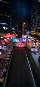 A busy urban street scene at night with heavy traffic, flanked by high-rise buildings adorned with illuminated signs. Bright red and white car lights create a vivid contrast against the dark surroundings. Overhead signs with colorful letters and numbers guide drivers to various parking options.