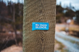 A blue sticker with the text 'No more SILENCE.' is affixed to a wooden post, set against a blurred natural background of trees and a pathway.