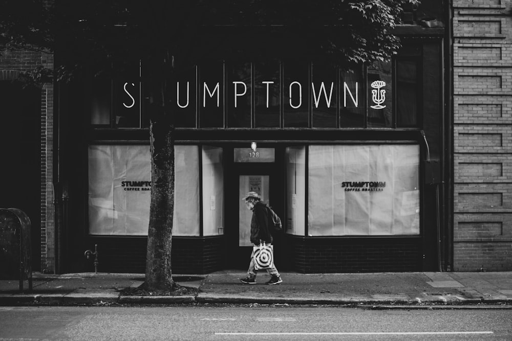 grayscale photo of woman in black coat walking on sidewalk