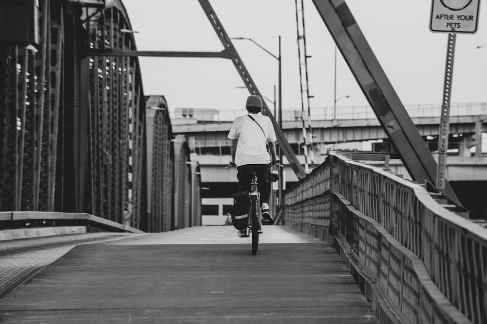 homme en chemise à manches longues blanche faisant du vélo sur le pont en niveaux de gris photographie