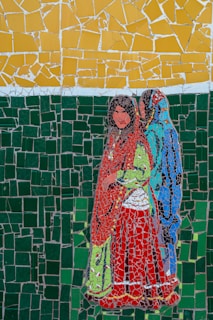 A mosaic artwork featuring two women dressed in traditional clothing. The background consists of shattered mosaic tiles in green and yellow, while the women’s attire is comprised of vibrant colors including red, green, blue, and orange. The mosaic tiles are arranged in a way that forms a detailed and colorful depiction.