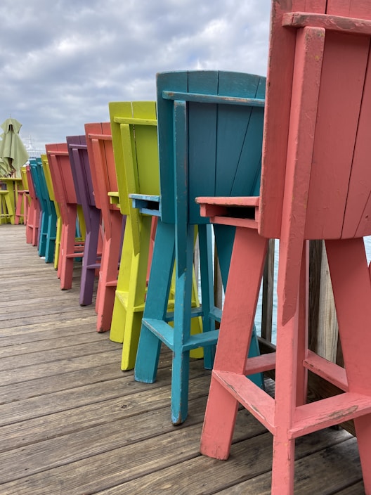 A row of colorful, tall wooden chairs lined up on a wooden deck, featuring vibrant shades of red, orange, yellow, green, blue, and purple. The sky is overcast, creating a muted backdrop that contrasts with the bright colors of the chairs.