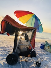A beach cart covered with a striped towel and shaded by a colorful rainbow umbrella near the shoreline during sunset. The sunlight creates a warm glow, while a cooler and beach bag sit nearby on the sand.