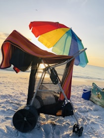 A beach cart covered with a striped towel and shaded by a colorful rainbow umbrella near the shoreline during sunset. The sunlight creates a warm glow, while a cooler and beach bag sit nearby on the sand.
