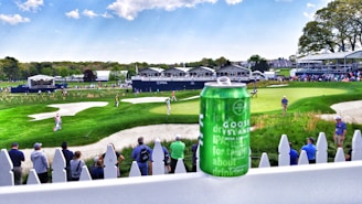 A vibrant golf course with lush greenery and a few sand traps is seen beyond a spectator area lined with a white picket fence. In the foreground, there is a green Goose Island IPA beer can on the railing. Several people are gathered along the fence, watching golfers playing in the background. Some tents and buildings are visible in the distance, likely part of a golf tournament setting.
