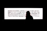 A silhouette of a person stands in front of a rectangular illuminated panel displaying numerous small images arranged in rows. The room is mostly dark, highlighting the luminous panel.